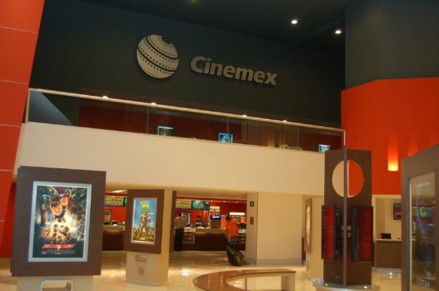 Eine Gruppe von Menschen in einem Cinemex-Kino in einem Einkaufszentrum, mit einer Person auf einem Stuhl, Tischen, Fotorahmen an der Wand, einem Schild, Deckenlampen und Geräten auf den Tischen.