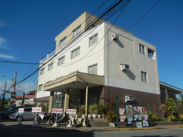Wohngebäude mit geparkten Autos, Schildern, Strommasten, Bäumen und einem bewölkten Himmel.