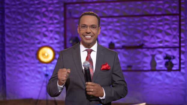 Stephen Colbert, der einen Anzug, Krawatte und Brille trägt, steht auf einer Bühne und hält ein Mikrofon in der Hand, mit einer Wand, einem Ständer, einer Lampe und verschiedenen Gegenständen dahinter.