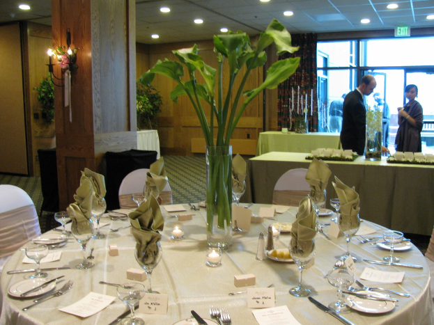 Ein Tisch mit weißen Tellern, Besteck und einem Glas, geschmückt mit Zweigen, umgeben von Stühlen, mit einem grünen Tisch dahinter; ein Mann in einem schwarzen Blazer und eine Frau in der Nähe einer Tür mit zwei Männern dahinter.