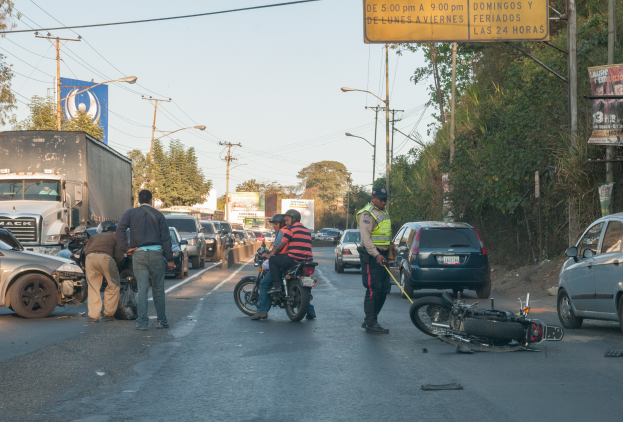 Gruppe von Menschen um ein verunglücktes Motorrad auf der Straßenseite mit mehreren Fahrzeugen, darunter ein Lastwagen, im Hintergrund und Bäumen, Masten, Lichter, Schilder und Himmel.