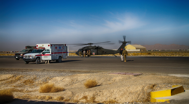 Ein Hubschrauber neben einem Rettungswagen auf einer grasbewachsenen Straßenseite mit Menschen in der Nähe, vor Höhen und einem klaren blauen Himmel.