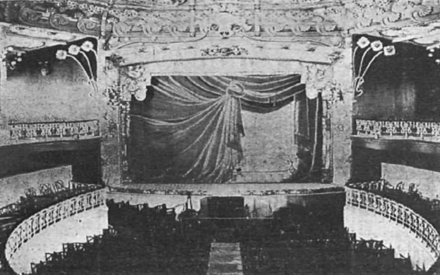 Schwarzes und weißes Foto des Auditoriums des Theatre Royal in London, das eine Bühne mit Vorhang, Geländer und ornamentierte Wandgestaltung zeigt, mit einigen sitzenden Besuchern und Text unten.