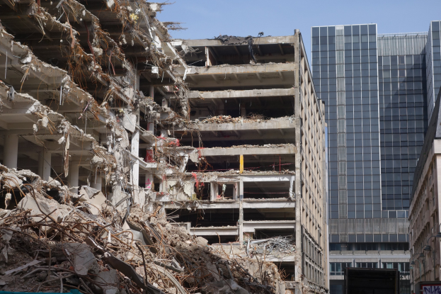 Großes Gebäude in der City-Mitte abgerissen, umgeben von intakten Gebäuden, Straßeninfrastruktur, Fahrzeugen, Bäumen und einem klaren blauen Himmel.