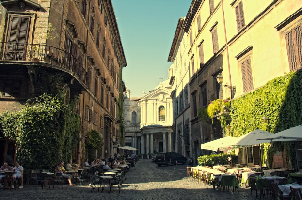Ein Weg, gesäumt von Tischen, Stühlen und parkenden Autos, flankiert von Gebäuden mit Pflanzen im Vordergrund und Himmel darüber.