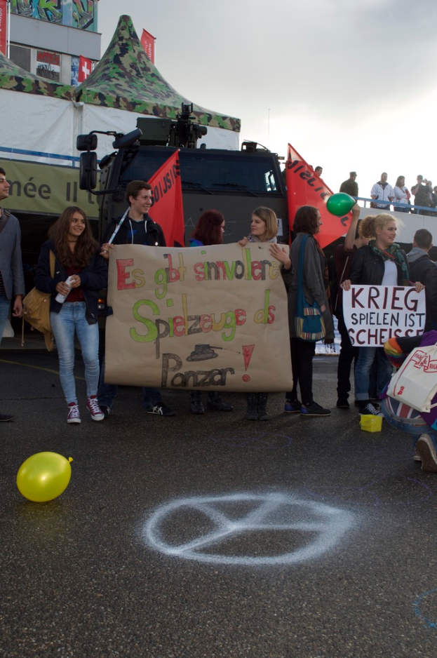 Eine Straßenszene in einer Stadt mit einem Ballon im Vordergrund, Menschen mit Schildern und Fahnen in der Mitte, einem Gebäude und einem Zelt im Hintergrund, unter einem bewölkten Himmel.