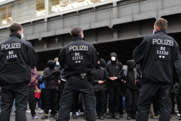 Polizisten in schwarzer Uniform und Masken stehen vor einer Menge während einer Outdoor-Demonstration, mit einer Brücke und einem Gebäude im Hintergrund.