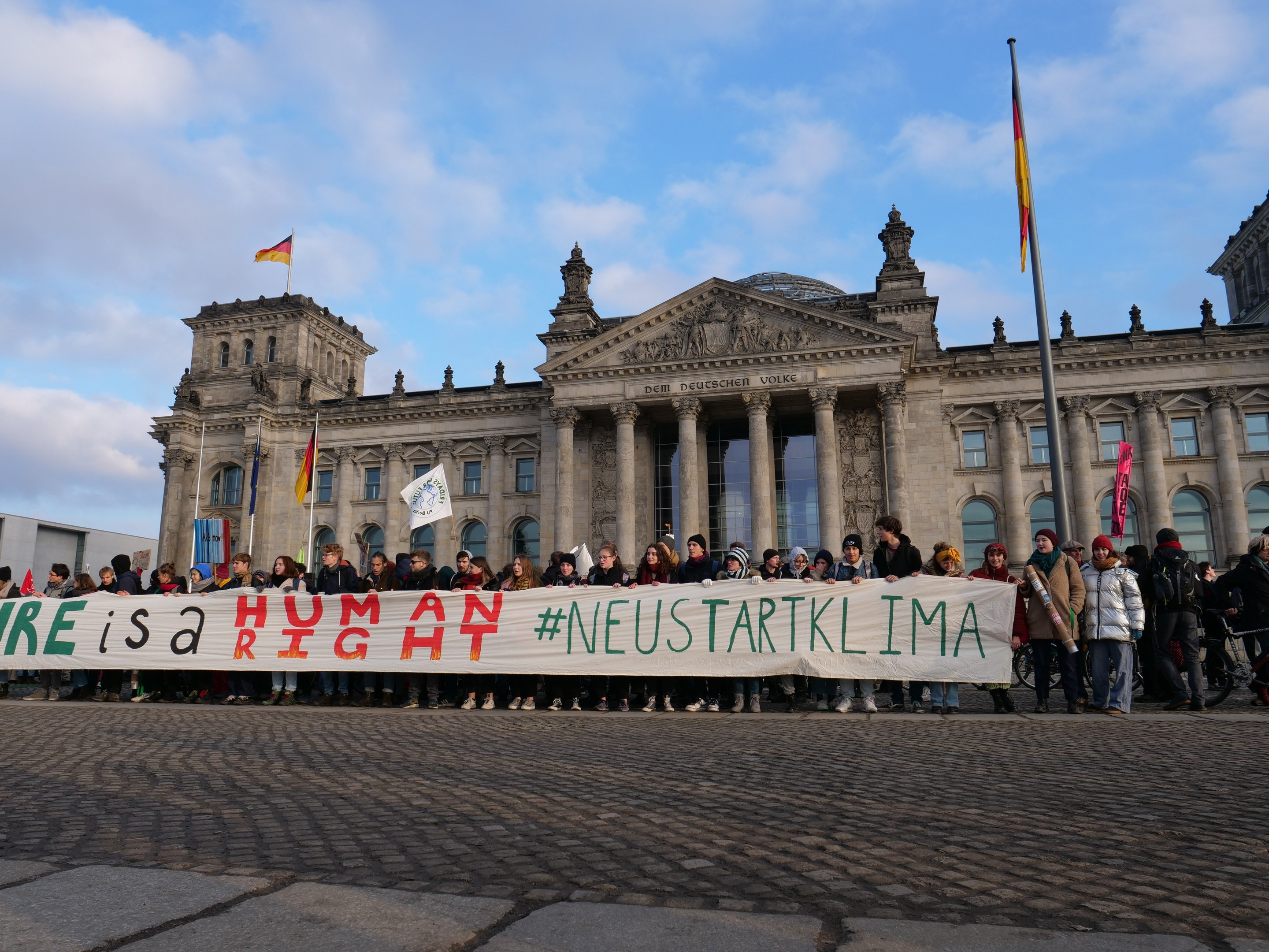 Gruppe von Menschen, die vor dem Reichstag in Berlin stehen und eine Fahne mit der Aufschrift "Wir sind ein Menschenrecht" halten.
