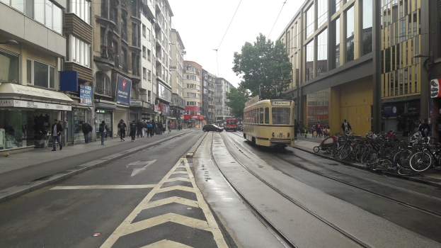 Eine Stadtstraße mit einer gelben Tram, Gebäuden mit Fenstern, Schildern, Passanten, parkenden Fahrrädern, einer Straßenlaterne, Oberleitungen, einem Baum und einem bewölkten Himmel.