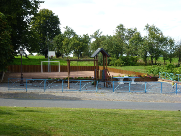 Ein Park mit einem Spielplatz in der Mitte, umgeben von einer Stra├če, Gras, Pflanzen, einem Zaun, einem Basketballkorb und B├Ąumen unter einem klaren blauen Himmel.