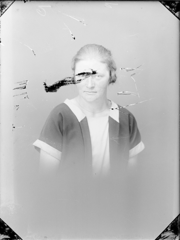 Schwarz-weiß-Foto einer Frau mit einem zerbrochenen Glasfenster im Hintergrund, die direkt in die Kamera schaut und mit leicht geöffneten Lippen und gefalteten Händen vor sich steht.