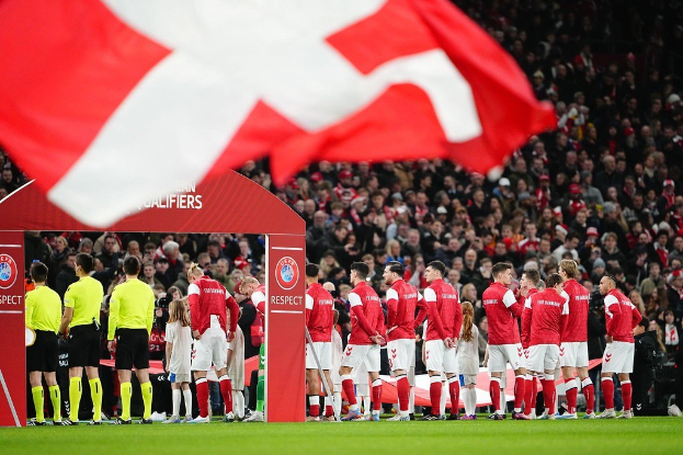 Gruppe von Menschen auf einem Fußballfeld mit einer roten und weißen Flagge im Vordergrund, einem Bogen mit dem Text "Bayern Munich vs Bayern Munich Betting Tips & Preview" im Hintergrund und einer großen Menge im Stadion.