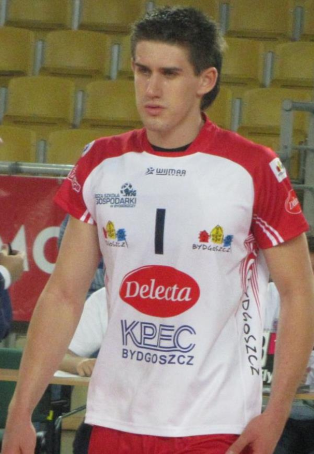 Ein Mann in einem roten und weißen Handballtrikot steht auf einem Feld und schaut entschlossen, mit Zuschauern und einer roten Fahne mit der Aufschrift "Krasnoyarsk vs Bydgoszcz" im Hintergrund.