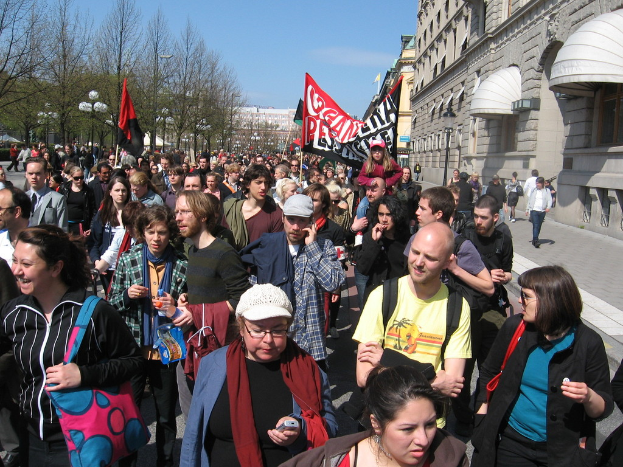 Eine Gruppe von Menschen, die auf einer Stra├če außerhalb der Stadt gehen, mit einem Mann in der Mitte, der eine rote und schwarze Fahne mit Text h├Ąlt, Geb├Ąude, B├Ąume, Stra├čenlaternen und eine rote Fahne im Hintergrund unter einem klaren Himmel.