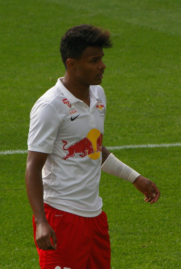 Ein Mann in einem weißen Hemd und roten Shorts steht auf einem Fußballfeld und schaut rechts mit entschlossener Miene, wobei er wie ein Flügelstürmer aussieht.