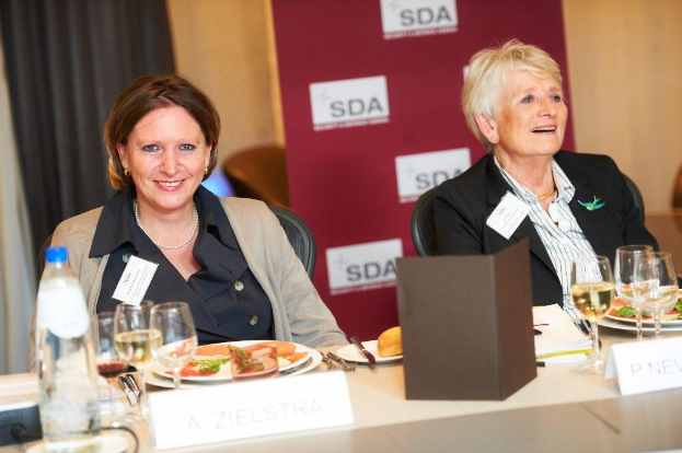 Zwei Frauen an einem Tisch mit Essen, Getränken und verschiedenen Gegenständen, die lächeln, mit einem Banner und Vorhängen im Hintergrund.