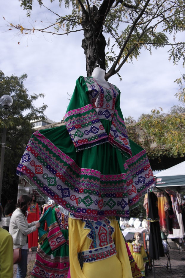 Eine Schaufensterpuppe steht in der Mitte eines belebten Einkaufsgebiets mit mehreren Geschäften, eine Person, die Kleider an einem Stab arrangiert, ein Gebäude und Bäume im Hintergrund.