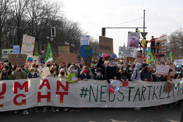 Eine große Gruppe von Menschen marschiert auf einer Straße und hält ein 'Menschliche Rechte'-Schild hoch, wobei einige Masken tragen und Schilder halten, unter einem klaren blauen Himmel mit Bäumen und Laternen im Hintergrund.