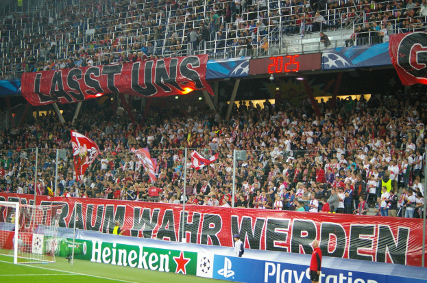 Fußballspiel in einem Stadion mit großer Zuschauermenge, saftig grünem Rasen, Torpfosten links, Bodenbanner, Menschen mit Fahnen und Hintergrundgeländer, Tafeln, Lichter und Gegenstände.