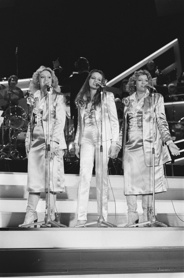 Drei Frauen singen in Microphone auf der Bühne mit Musikern im Hintergrund, schwarz-weiß.