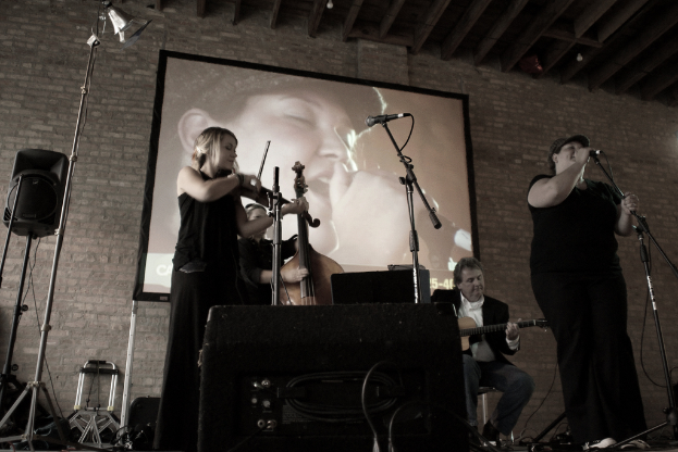 Eine Gruppe von Musikern in Schwarz, mit einem Mädchen, das Geige spielt, zwei Gitarristen im Hintergrund und einer Frau, die rechts singt.