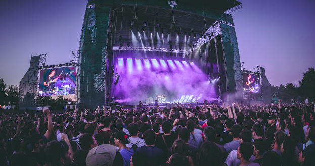 Eine große Menschenmenge auf einem Musikfestival, mit Musikern auf einer zentralen Bühne, die von Scheinwerfern beleuchtet wird und von Bildschirmen umgeben ist, unter einem klaren blauen Himmel mit einem Gebäude und Bäumen im Hintergrund.