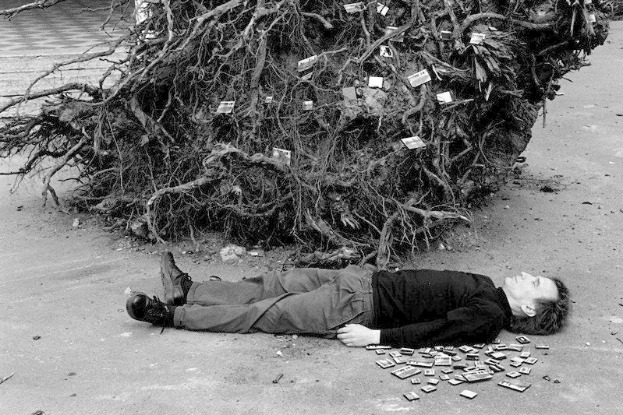 Ein Mann liegt neben einem Geldhaufen auf dem Boden, trägt Wanderschuhe und hat einen Baum im Hintergrund, auf einem Schwarz-Weiß-Bild.