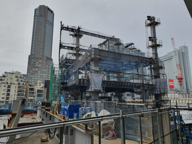 Großer Baustelle in einer Stadt mit einem Metallzaun, Gebäuden, Masten, einer Plane, Fahrzeugen, Käble, und einem bewölktem Himmel, beschriftet als neuer Bau einer Kraftwerk.