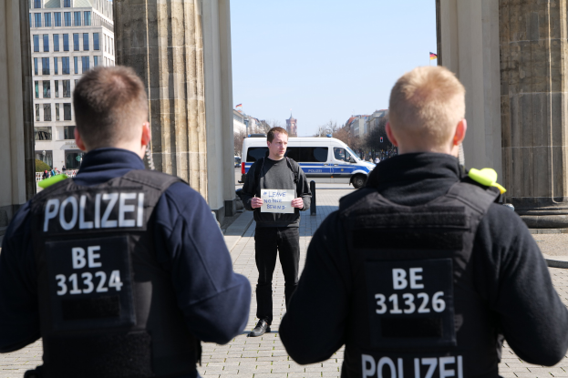 Drei Polizeibeamte stehen vor einem Gebäude, mit einem Mann, der ein Papier zwischen ihnen hält, flankiert von Säulen und einer belebten städtischen Umgebung.