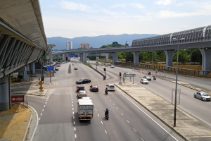 Eine vielbefahrene Autobahn mit mehreren Fahrspuren, neben einer Brücke, mit Straßenlaternen, Verkehrsampeln, Schildern, Bäumen, Gebäuden, Hügeln und einem bewölkten Himmel.