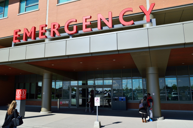 Gruppe von Menschen vor einem Notdienstgebäude mit Glaswänden, Säulen und Beschilderung, einschließlich eines Mülleimers.