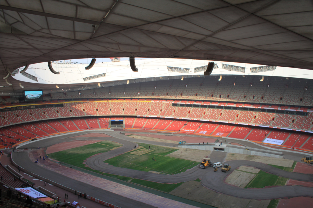 Großes Stadion voller Zuschauer, Fahrzeuge auf der Straße, grüner Boden, Texttafeln, Geländer und ein Bildschirm, mit Deckenbeleuchtung darüber.