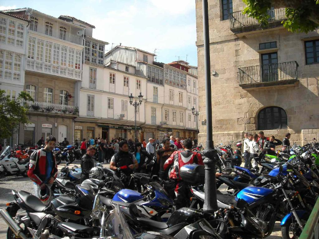 Ein cremefarbenes Gebäude mit zahlreichen Fenstern, das an ein Backsteingebäude mit einem Metallbalkon angrenzt, mit mehreren Fahrrädern davor geparkt und Menschen, die stehen und sich unterhalten.