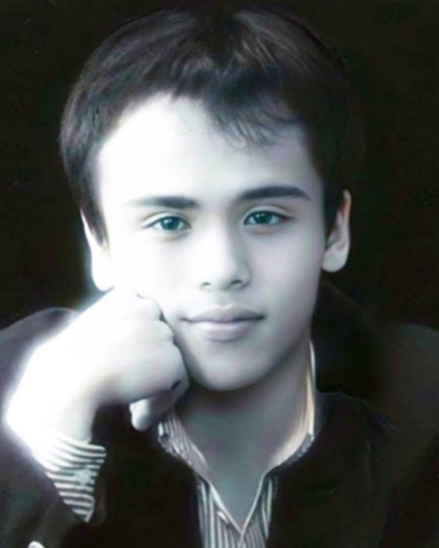 Ein junger Mann in einer schwarzen Jacke posiert selbstbewusst vor einem dunklen Hintergrund mit einem entschlossenen Gesichtsausdruck.