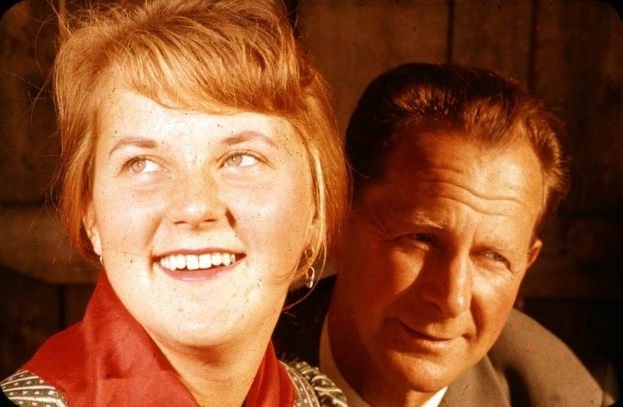 Zwei Menschen in inniger Umarmung, wobei der Mann seinen Arm um die Taille der Frau legt und ihr Kopf auf seiner Schulter ruht, vor einer schmucklosen Wand stehend.