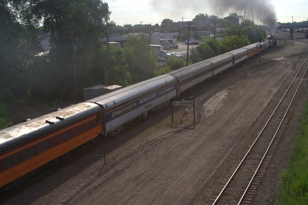 Ein Zug auf Schienen mit Bäumen, Strommasten mit Kabeln, Autos und Gebäuden links, eine Brücke vorne und Rauch, der aus dem Zug austritt.