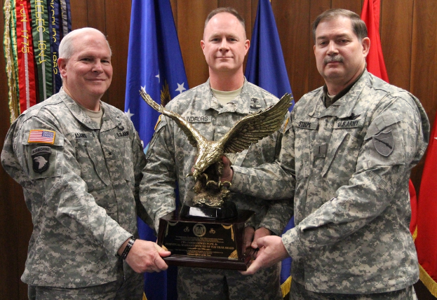 Drei Männer in Uniformen stehen mit Auszeichnungen, Flaggen dahinter und buntem Text in der linken oberen Ecke.