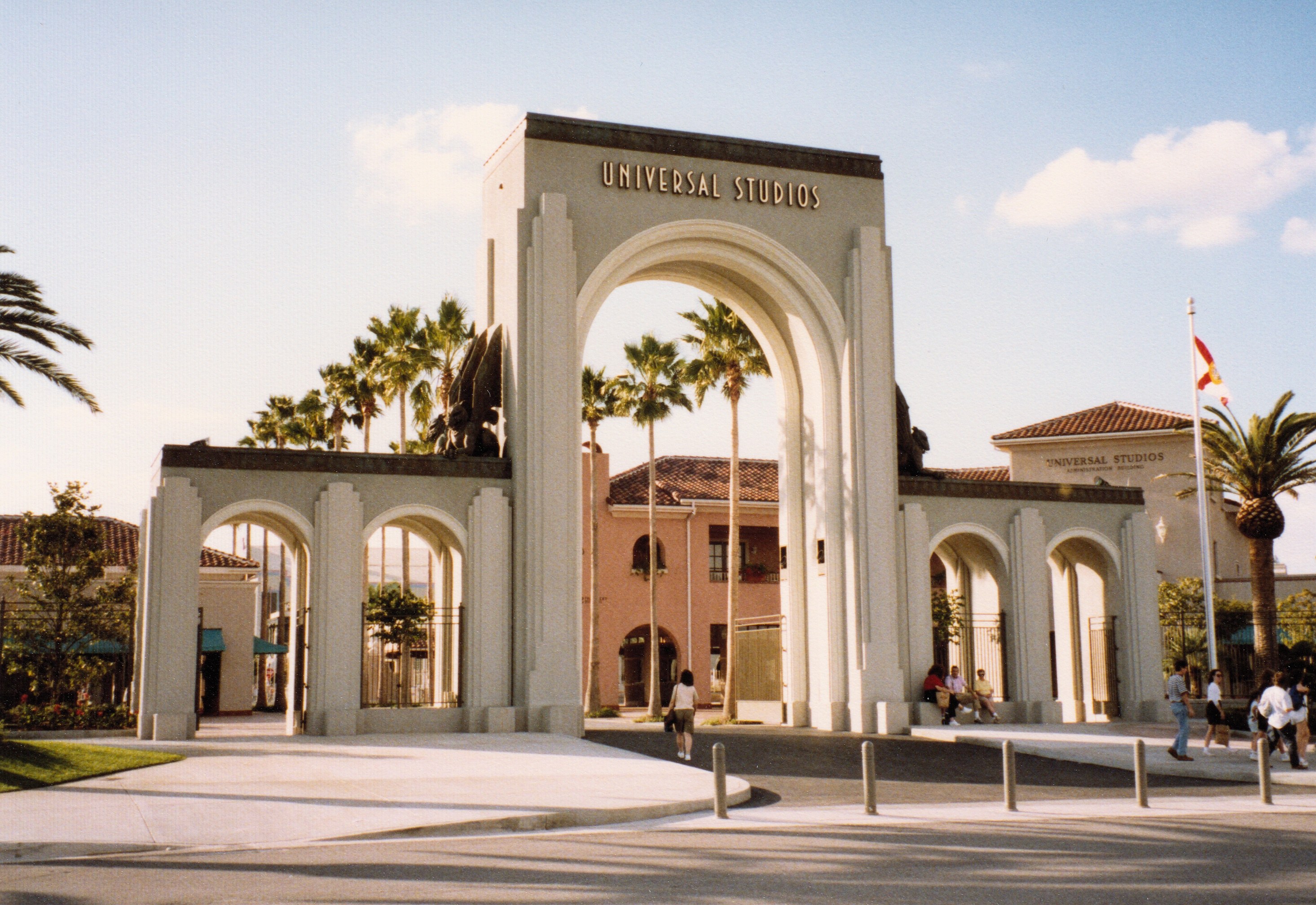 Eingang zu Universal Studios Hollywood mit einem Bogen, einer Statue, Menschen, Bäumen, Gebäuden, einer Fahnenstange und einem bewölkten Himmel.