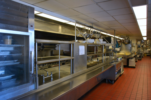 Gewerbeküche mit Edelstahl-Theke, Deckenbeleuchtung, hängenden Utensilien, Waschbecken, Wasserhähnen, Schränken und diversen Gegenständen auf Regalen.