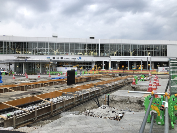 Baustelle am Toyama-Bahnhof mit einem großen Gebäude im Bau, umgeben von Fahrzeugen, Verkehrskegeln, Pfählen und Schildern im Vordergrund unter einem bewölkten Himmel.