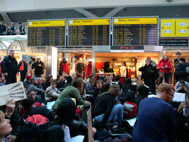 Eine große Gruppe von Menschen sitzt und steht in einem Flughafen-Terminal während einer Protestaktion, mit Gepäck und Papieren, während Anzeigetafeln, Schaufensterpuppen in Kleidern und Deckenbeleuchtung im Hintergrund zu sehen sind.