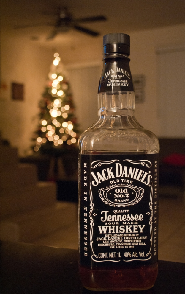 Eine Glasflasche mit der Aufschrift 'Jack Daniel's' steht auf einem Tisch vor einem beleuchteten Weihnachtsbaum, mit einem Ventilator darüber und einer weißen Wand im Hintergrund.