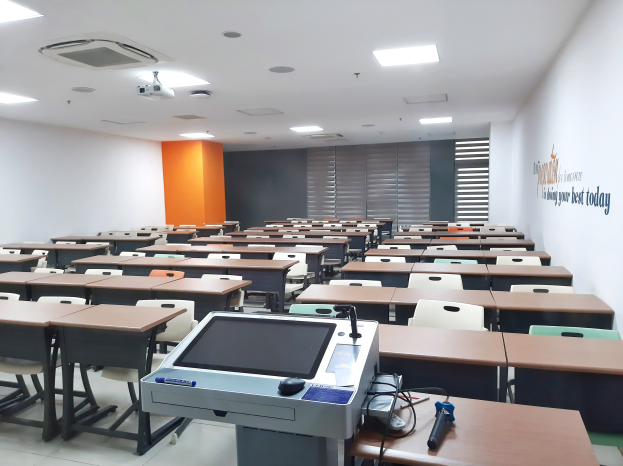 Ein Klassenzimmer mit Tischen und Stühlen in Reihen, ein deckengehängter Projektor und Gegenstände auf den Tischen, mit Text an der rechten Wand, der auf eine Privatschule in Singapur hinweist.