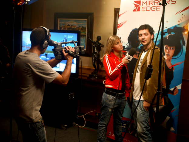 Ein Mann mit einer Videokamera und Headset filmt ein Interview zwischen einem Mann und einer Frau, mit einem Fernseher und einem gerahmten Foto an der Wand dahinter.