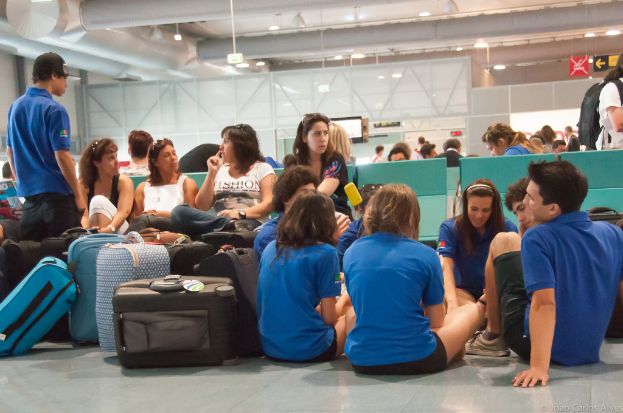 Ein belebter Flughafeninnenraum mit vielen Menschen, die auf Flüge warten, Gepäck auf der linken Seite und gläserne Türen mit Richtungsschildern im Hintergrund.