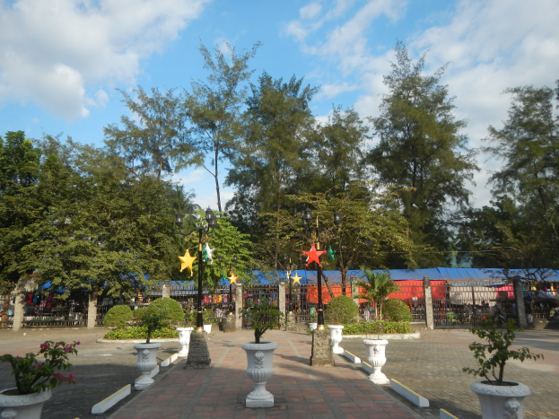 Ein Park mit Topfpflanzen und Bäumen in der Mitte, umgeben von Straßenlaternen, Schmuckelementen, Grills und einer bewölkten Himmel im Hintergrund.