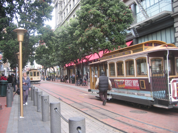 Ein Zug auf Schienen mit Text an der Seite, eine Person in der Nähe, kleine Pfosten mit Seilen links, Straßenlaternen, Bäume, viele Menschen im Hintergrund, ein Müllcontainer links und Gebäude mit Leitern rechts.