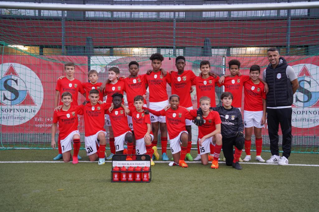 Eine Gruppe junger Männer in roten T-Shirts und schwarzen Shorts steht auf einem Fußballfeld vor einer Kiste roter Gegenstände, im Hintergrund Schilder mit 'FC Red Black Eglaute - Champions League'