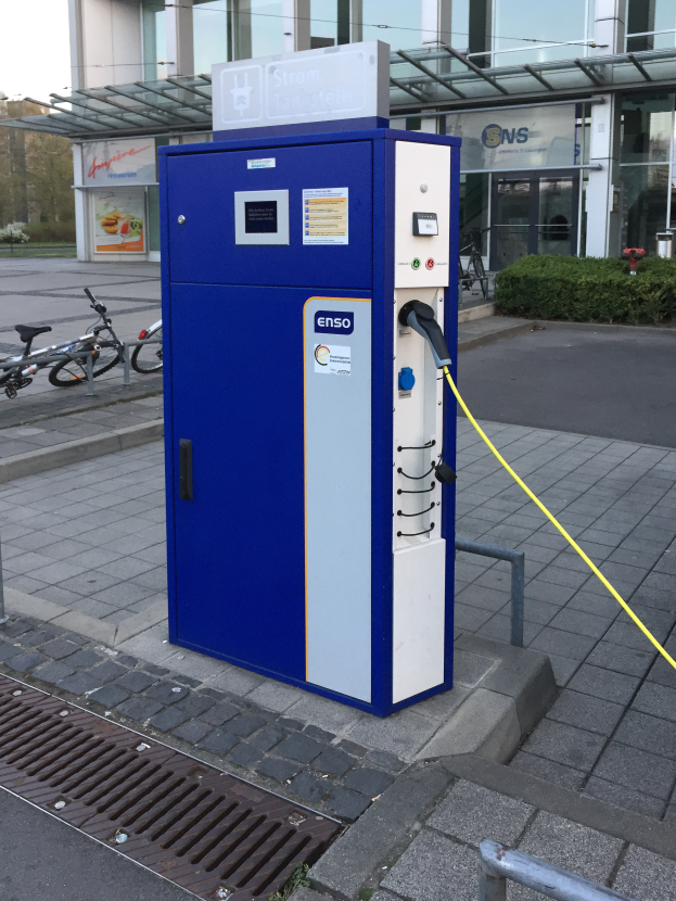 Eine Ladestation für Elektrofahrzeuge mit einer blauen und weißen Maschine im Vordergrund, mit Fahrrädern, Pflanzen, einem Gebäude, Bäumen und einem klaren blauen Himmel im Hintergrund.