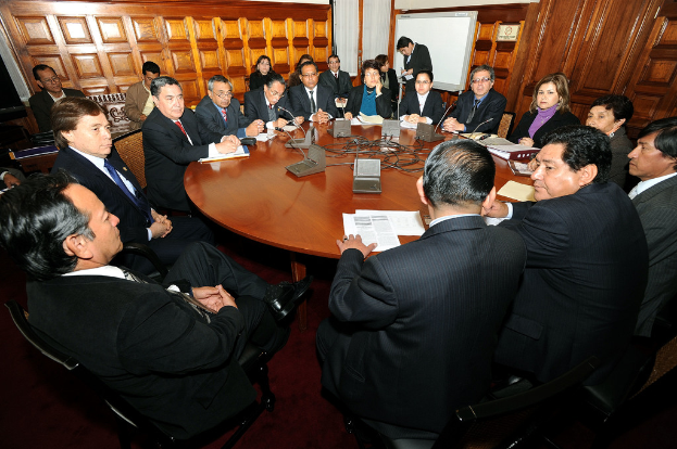 Rundtischgespräch mit Delegierten, die diskutieren, Mikrofone, Papiere, Kabel und Dateien auf dem Tisch sowie eine hölzerne Wand im Hintergrund.